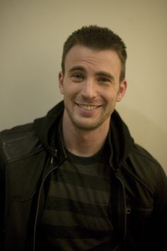 a man in a black jacket smiling at the camera with a white wall behind him