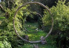 a circular sculpture made out of branches in the middle of some grass and trees with rocks around it