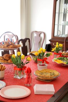 a red table topped with plates and bowls filled with food next to a vase full of yellow tulips