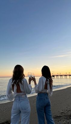 two women standing on top of a beach holding wine glasses in front of the ocean