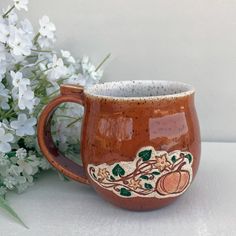 a ceramic mug with an autumn scene on it next to white flowers and greenery