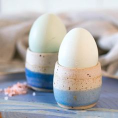 two eggs are sitting in small cups on a blue tableclothed surface, one is white and the other is blue with speckles