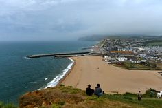 two people sitting on the edge of a cliff looking out at an ocean and town