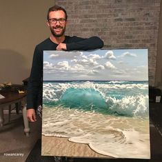 a man standing in front of a painting holding up a large blue wave on the beach