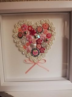 a heart made out of buttons and pearls in a white frame with pink ribbon on the bottom