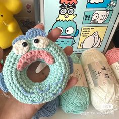 a hand holding a crocheted donut with eyes and mouth in front of yarn balls