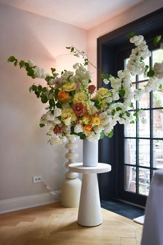 two white vases filled with flowers on top of a wooden table next to a window