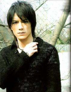 a young man with black hair and piercings standing in front of a tree wearing a sweater