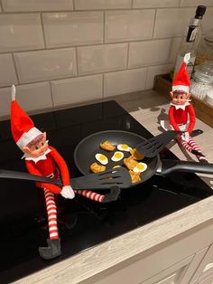 two elfs are sitting on the kitchen counter and one is holding a frying pan with eggs in it