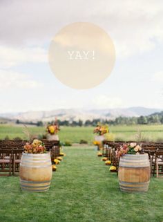 an outdoor ceremony set up with wooden barrels and flower arrangements on the grass in front of rows of chairs