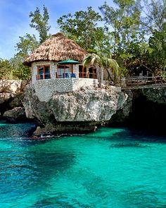 a house built into the side of a cliff in the ocean with blue water and green vegetation