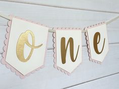 a pink and gold one banner hanging from a string on a white wooden wall with the word one spelled out