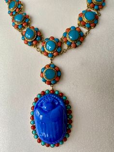 A vintage striking and beautiful  1930's Egyptian Revival coral and turquoise glass Scarab drop necklace. A single row of turquoise and coral glass cabochon rosettes, 15 in total, with a pendant of a further rosette and a large turquoise glass Scarab Beetle with a surround of turquoise and coral glass cabochons. The metal mounts are gilt metal and the cabochons collet set. A lovely clasp set with 3 glass turquoise cabochons, hook and ring clasp secures. Length of 17 inch with 4.1/2 inch drop. In Traditional Blue Cabochon Necklace, Traditional Blue Cabochon Necklaces, Antique Blue Cabochon Necklace, Antique Blue Medallion Necklace, Egyptian Revival Jewelry, Czech Jewelry, Czech Glass Necklace, Exotic Jewelry, Scarab Beetle