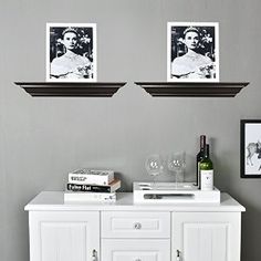 two framed pictures hang above a white cabinet with wine glasses and books on the top