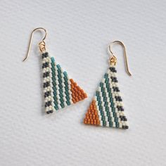 two triangle shaped beaded earrings with gold hooks on white tablecloths, one has an orange and green striped design