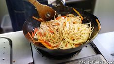 a wok filled with noodles and carrots on top of a gas burner