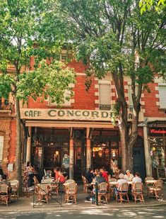 people sitting at tables in front of a cafe on the street corner with trees lining the sidewalk