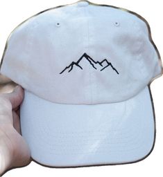 a person holding up a white hat with mountains embroidered on the front and side of it