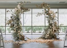 two white floral arrangements on the floor in front of large windows at an indoor wedding ceremony