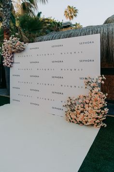 a sign with flowers on it sitting in front of a palm tree and thatched roof