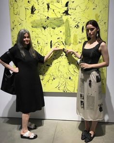 two women standing next to each other in front of a large yellow piece of art