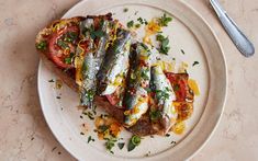 a white plate topped with two pieces of toast covered in fish and vegetables next to a fork