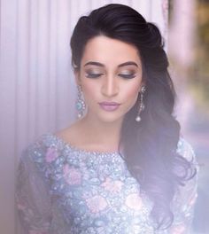 a woman with long dark hair wearing a blue dress and earrings, standing in front of a white wall