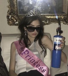 a woman sitting on top of a couch holding a blue bottle and wearing a pink sash