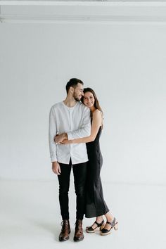 a man and woman standing next to each other with their arms around each other in front of a white wall