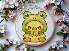 a cross stitch frog sitting on top of a flowery branch in front of some white flowers