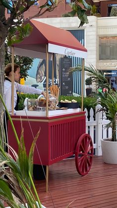 a woman standing behind a red cart with food on it's side and palm trees in the background