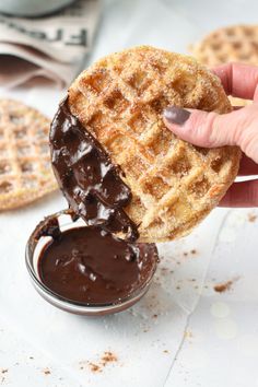 a hand holding a waffle with chocolate sauce on it and some cookies in the background