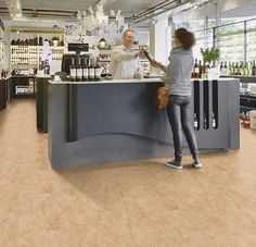 a woman standing at the counter in a store with bottles on shelves behind her and another person holding a bag