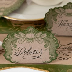 two place cards are sitting on top of each other, with gold rimmed plates in the background