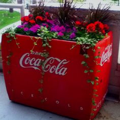 an old coca - cola cooler with flowers growing out of it's sides on the porch