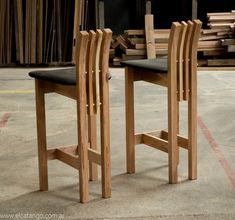 two wooden chairs sitting next to each other on top of a cement floor in front of stacks of wood