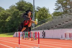 a woman in a graduation gown jumping over a hurdle on a track with her arms outstretched