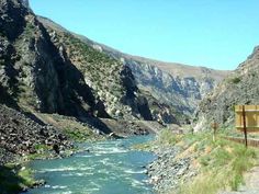 a train traveling down tracks next to a river and mountain range in the distance with mountains on either side