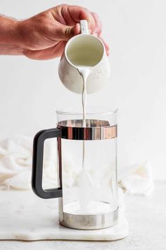 someone pours milk into a glass coffee pot from a metal mug on a white surface