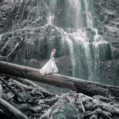 a woman standing on a log in front of a waterfall wearing a long white dress