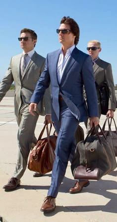 a man in a suit and sunglasses walking with two other men behind him carrying luggage