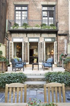 an outdoor living area with chairs and tables in front of a brick building, surrounded by greenery