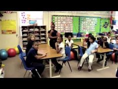 a group of children sitting at desks in a classroom with adults standing around them