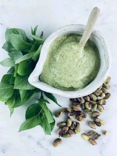 a white bowl filled with green dip surrounded by pistachio seeds and leaves on a marble surface