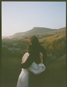 two people embracing each other in the middle of a grassy field with hills in the background