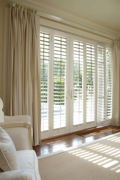 a living room with white furniture and large windows covered in plantation shutters, sun shining through the blinds