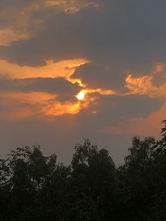 the sun is setting over some trees in the distance, with clouds above and behind it