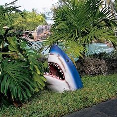 a fake shark with its mouth open sitting in the grass next to some trees and bushes