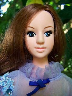 a doll with brown hair and blue eyes wearing a purple dress in front of some trees