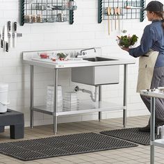 a woman standing in front of a kitchen sink with plants on the counter and shelving behind her